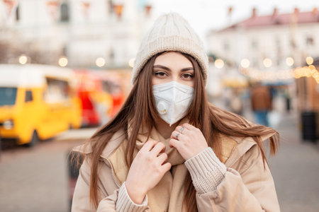 Fashionable portrait of a pretty young woman with a medical protective mask in fashionable outerwear with a knitted hat and jacket walks in the cityの写真素材