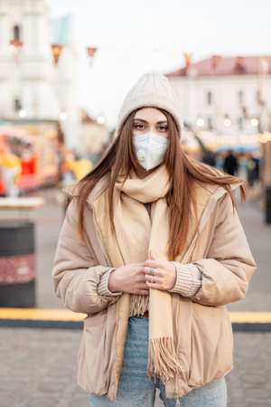 Modern portrait of a pretty young girl in a medical mask and a stylish outwear with a knitted hat, jacket and scarf walking in the city. female style and beautyの写真素材