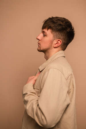 Handsome young man with hairstyle in fashionable clothes with beige jacket stands on a beige...