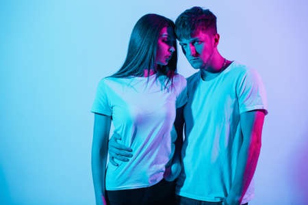 Stylish beautiful young couple in white t-shirts with neon and pink light in the studio. Pretty girl model and handsome man with colored backgroundの写真素材