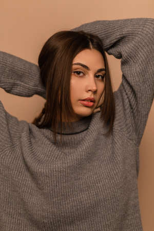 Fashionable portrait of a pretty young girl model in a stylish gray knitted sweater posing and looking at the camera on a beige background. beauty girl indoorsの写真素材