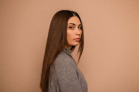 Natural beautiful portrait of a fashionable sensual girl with brunette hair in a fashionable knitted gray sweater on a beige background. Woman in comfortable clothes in studioの写真素材