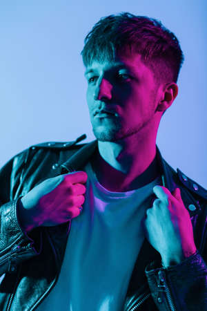 Colored fashion portrait of a handsome stylish guy with hairstyle in trendy outwear with vintage leather jacket on neon pink and purple lightの写真素材
