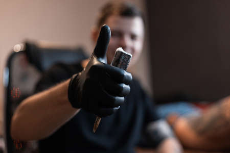professional tattoo artist man in black gloves holds a tattoo machine and shows thumbs up - like. Working process of creating a tattoo in the studioの写真素材