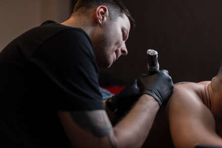Young professional tattoo artist man with a modern tattoo machine in a working process makes a...