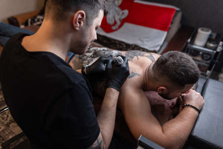 Man professional tattoo artist in black gloves makes a tattoo on the male back in the studio. creative work processの写真素材