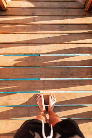 man in shorts stands on a wooden bridge near the sea. Men's feet on a wooden pier at sunset, top view. Summer rest concept.の写真素材