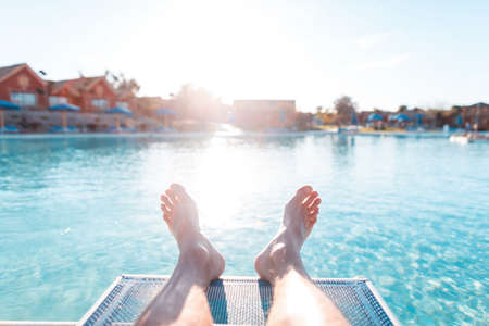 young man lies and rest on beach lounger near the pool at sunset. Man's feet on a background of blue water and hotel bungalow houses. First person view relaxの写真素材