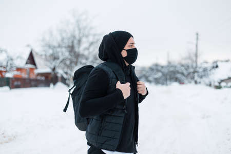 Handsome young fashion man with protective black mask in trendy black clothes with a backpack travels in winter season with snow in town. Coronavirus and styleの写真素材