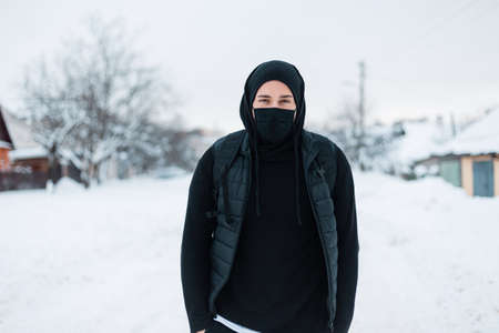 handsome young stylish hipster man with a protective mask in black fashionable outerwear with a hoodie, vest, hat and backpack walks on a winter day with snow. Guy in the town. coronavirus conceptの写真素材