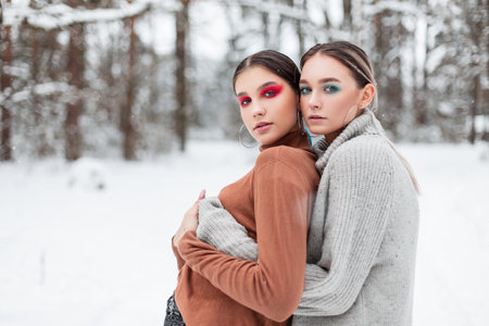 Two beautiful young women with bright creative makeup in a vintage knitted sweater hugging outdoors in the forest with snowの写真素材