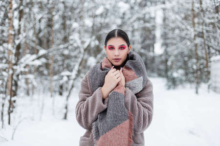 fashionable vogue girl with bright colored makeup in stylish winter clothes with a scarf and fur coat stands in the forest with snow and looks into the camera.の写真素材