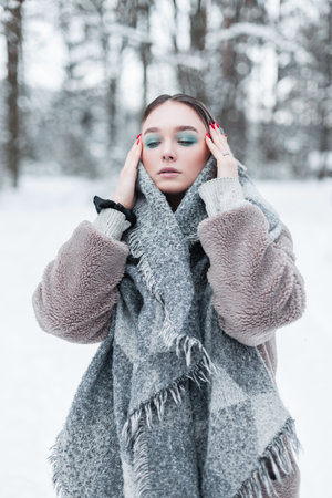 Stylish beautiful young russian girl model in winter clothes with a scarf and a warm fur coat in the winter snowy forestの写真素材