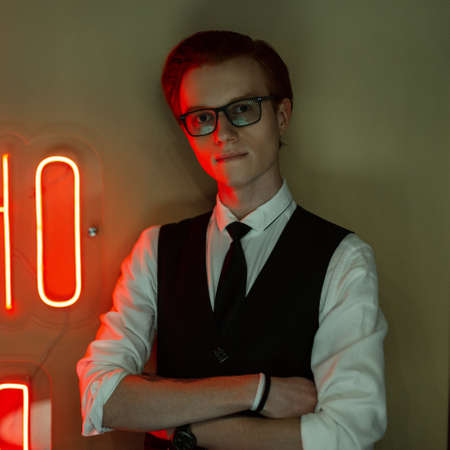 Business handsome young redhead man with glasses in fashionable clothes with a suit and tie stands near a green wall with red neon lightの写真素材