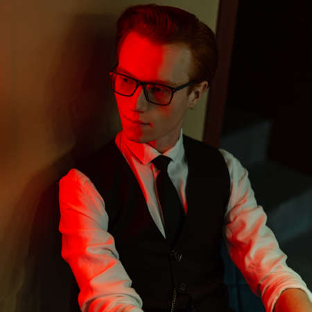 Stylish handsome young man student model with glasses in fashion business clothes with vest, shirt and tie sits on a chair near the wall with green and red neon lightsの写真素材