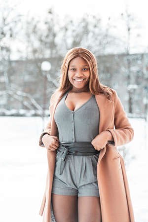 Sexy beautiful African young woman with smile in fashionable clothes with a beige coat correcting a jumpsuit on a white background with snow. Winter feminine style and beauty. black girl walking in townの写真素材