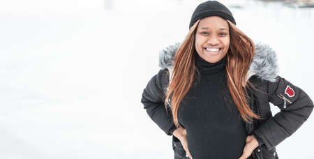 Beautiful young stylish black girl with a smile in fashionable winter clothes with a jacket and hat in the white snowの写真素材