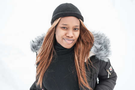 Stylish portrait of a beautiful young girl with black skin in a fashionable jacket, sweater and hat on a white background with snowの写真素材