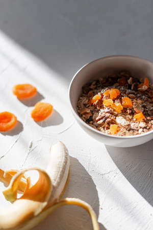 Delicious breakfast raw muesli with oatmeal, dried apricots, nuts and banana on a white table with early morning sun rays. Healthy food at sunrise. Start of the day and Proper Nutrition For Healthの写真素材