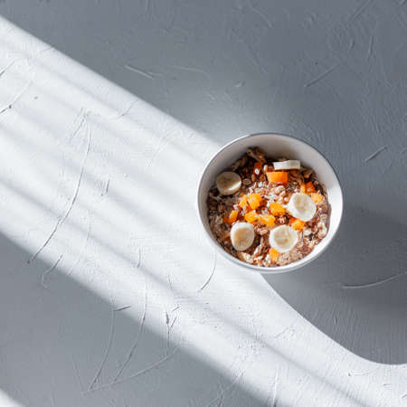 Healthy delicious breakfast muesli with oatmeal, dried apricots and banana in a white bowl on a white table on the rays of the sun in a morning, top viewの写真素材