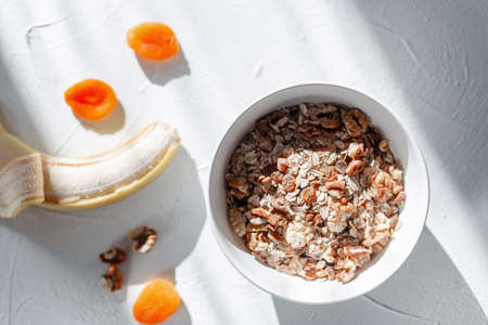 Healthy raw food muesli with oatmeal, dried apricots, walnuts and banana on a white table with early morning sunlight, top view. Vegan breakfastの写真素材