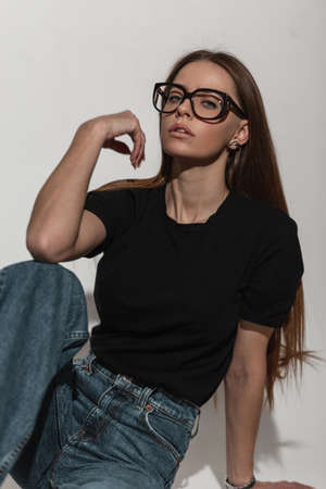Sexy glamorous beauty woman with cool glasses in fashion black shirt mock-up and jeans sits in studio on a white background. Pretty lovely girl in fashionable summer clothesの写真素材