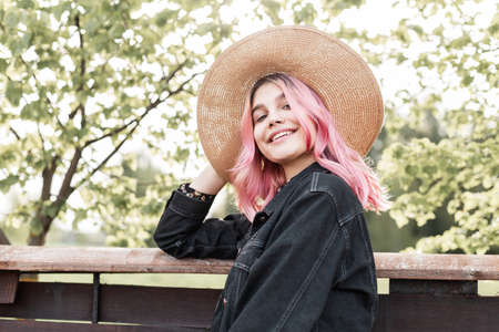 Smiling young woman with pink hair in straw hat in fashionable denim jacket in beautiful summer dress is resting near wooden fence in country. Cute fashion girl with smile enjoys weekend in nature.の写真素材