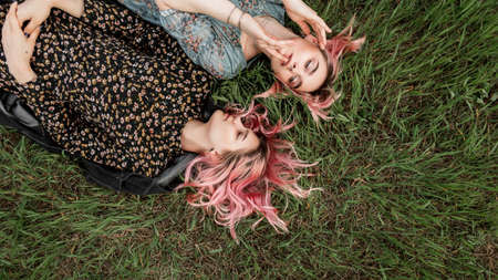Attractive girlfriends with beautiful pink hair in fashionable summer clothes rests on fresh green grass in field outside city. Fashion gorgeous girls in elegant summer dresses enjoy weekend on natureの写真素材
