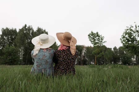 Rear view on fashionable young women with pink hair in elegant straw hats in trendy dresses with floral pattern from new collection on grass outside city. Stylish modern girls enjoy beauty of nature.の写真素材