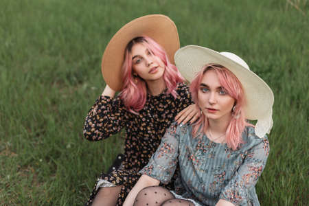 Two fashionable beautiful young women in trendy clothes with a straw hat sit on the grass and relax in the countrysideの写真素材