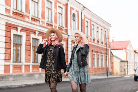 Smiling joyful young sister-twins with pink hair in fashionable summer youth clothes in stylish vintage straw hats hold hands and smiles outdoors. Happy girlfriends in trendy dresses walks on the road.の写真素材