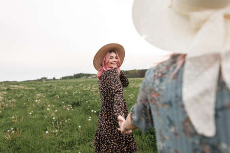 Trendy woman in fashion blue dress in hat holds hand of a positive girlfriend with pink hair in with vintage straw hat with charming smile. Cute sisters walk in the field with beautiful nature. Follow me.の写真素材
