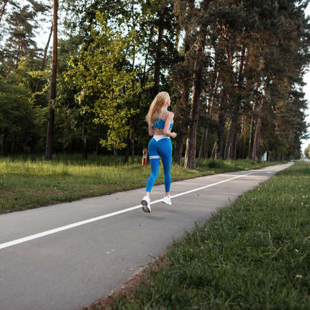 sportive beautiful young blonde girl in blue sportswear with white sneakers runs on the nature in the parkの写真素材