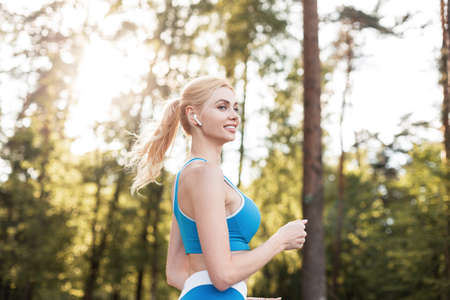 Happy attractive woman with lovely smile in sports blue stylish top with modern wireless headphones jogging in forest on bright sunny summer day. Joyful girl runs and listens music at sunsetの写真素材