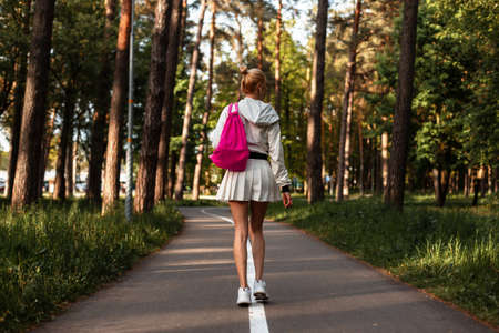 beautiful young sporty blonde girl in a fashionable jacket and a sexy sports skirt with a pink backpack walks in the park and goes to workoutの写真素材