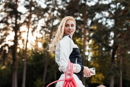 Smiling pretty happy young blonde in sporty stylish black-white jacket with pink trendy backpack poses at sunset in forest. Positive girl fashion model with smile in trendy sportswear walks in park.の写真素材