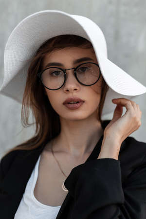 Beauty portrait young woman with clean skin in vintage glasses in black fashion jacket in straw white elegant summer hat near gray wall on street. elegance style.の写真素材