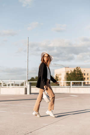Young European woman model in straw hat in black blazer in fashionable pants in casual sneakers with bag walk outdoors. Lovely stylish girl in beautiful beachwear in parking lot on sunny day.の写真素材