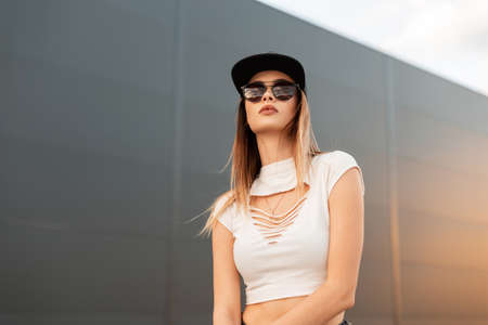 Fashionable portrait cool urban woman with sexy lips in white stylish torn top in jeans in dark trendy sunglasses and black baseball cap on gray background outdoors at sunset. summer youth fashion.の写真素材