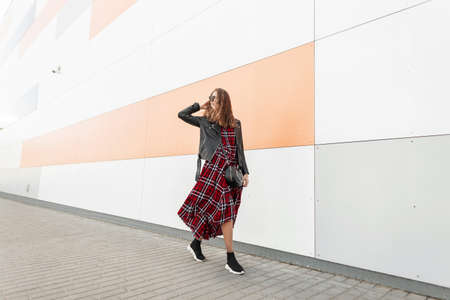 Fashion model happy urban hipster in dark sunglasses with smile in fashionable youth red-black outfit and stylish sneakers travel on city. Smiling american girl walks near modern wall on street.