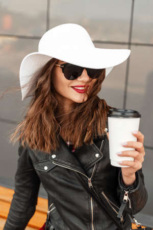 Beauty portrait smiling woman with cup of coffee with red lips in vintage hat in black fashion jacket in cool sunglasses near wall on street. Happy beautiful girl with pretty smileの写真素材