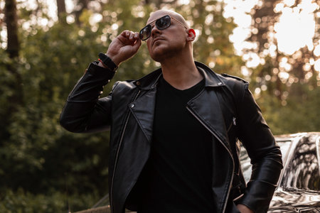 Fashion brutal man with beard in black vintage leather jacket in trendy sunglasses posing near modern car in forest at sunset. Cool bald guy travels by auto and rest on nature from bustle of city.の写真素材