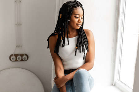 Gorgeous African American woman with cool dreadlocks in fashionable clothes sits near white wall and...