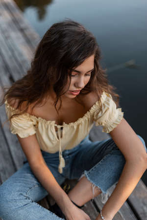 Beautiful young woman with curly hair in summer denim clothes with yellow top blouse and jeans sits on wooden pier by the lakeの写真素材