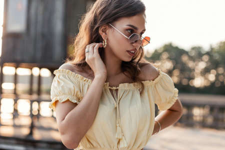 Stylish attractive girl in fashion sunglasses in a summer yellow outfit stands near the lake in...