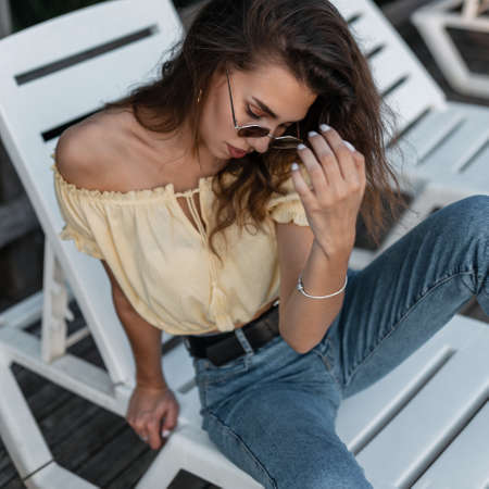 Fashionable portrait of trendy hipster woman with stylish sunglasses in yellow top blouse and blue vintage jeans sitting and rest on beach loungerの写真素材