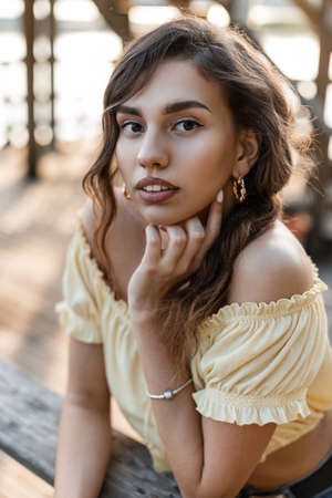 Summer portrait of a young beautiful woman model with curly hairstyle fashion yellow top blouse stands on pier near a lakeの写真素材