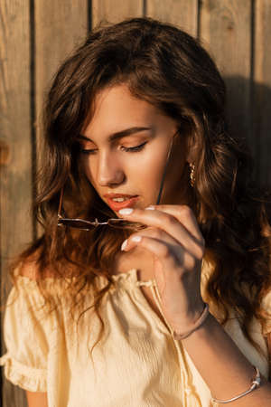 Fresh portrait of a young beauty hipster woman in a yellow blouse top put on a sunglasses near a wooden wall at sunsetの写真素材