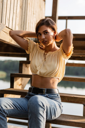 Young beautiful woman hipster in a yellow blouse top and blue jeans sits on wooden steps outdoorsの写真素材