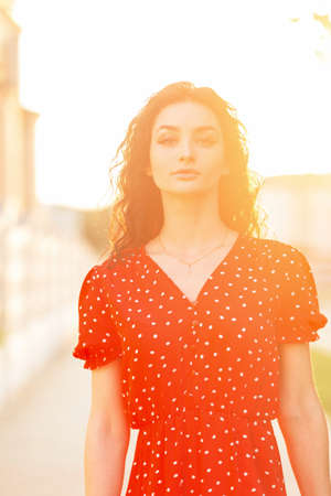 Summer fresh portrait of fashion woman with curly hair in a summer red dress walks at sunset outdoorsの写真素材